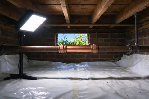 Short new section of copper pipe joined into an older copper line with two press couplings, under a crawl space floor